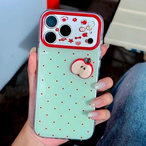 May include: A clear smartphone case with a light mint green back featuring small red polka dots. The camera area is framed in bright red and decorated with tiny red apple, flower, and star icons. A three-dimensional red apple charm is attached.