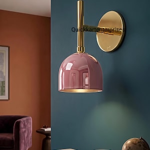 May include: A wall-mounted lamp featuring a polished gold-tone metal arm and a circular wall plate, paired with a glossy, dome-shaped dusty pink shade. The text Quickkartenterprises is visible on the gold arm of the lamp fixture.