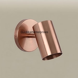 May include: A wall-mounted spotlight fixture with a brushed copper finish. The lamp features a circular base plate connected to a cylindrical head by a short arm. The text Quickkartenterprises is printed on the side of the cylindrical lamp head.