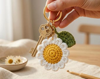 Peut inclure: Un porte-clés en métal doré avec une breloque marguerite crochetée au centre jaune et aux pétales blancs, accompagnée d'une petite feuille verte crochetée. Une clé en métal couleur laiton est fixée à l'anneau fendu circulaire.