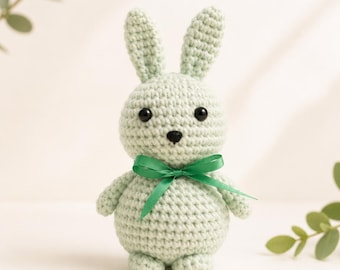 Peut inclure: Un lapin en crochet vert menthe clair avec deux longues oreilles droites, un corps rond et une petite tête. Le lapin possède deux yeux en perles noires et un nez brodé noir. Un ruban en satin vert foncé est noué en nœud autour du cou.