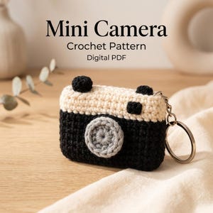 Puede incluir: Un llavero de ganchillo con forma de mini cámara con base negra, parte superior crema y lente circular gris. El texto Mini Camera Crochet Pattern Digital PDF aparece sobre el objeto, que cuenta con una anilla de metal plateado.