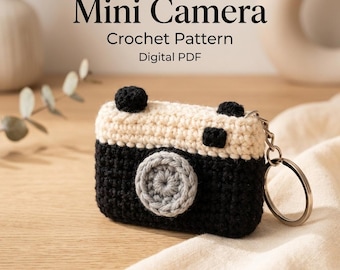 Peut inclure: Un porte-clés au crochet en forme de mini appareil photo avec une base noire, un haut crème et un objectif circulaire gris. Le texte Mini Camera Crochet Pattern Digital PDF est affiché au-dessus de l'objet, doté d'un anneau en métal argenté.