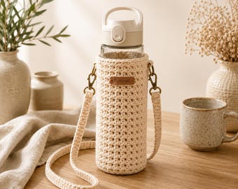 Peut inclure: Une bouteille d'eau en verre transparent avec un couvercle en plastique blanc et une housse en crochet couleur crème. La housse comporte une petite étiquette en cuir marron avec le mot Handmade et une bandoulière en crochet assortie.