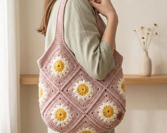 Peut inclure: Un sac fourre-tout en crochet composé de motifs carrés avec un motif de marguerite aux centres jaunes et pétales blancs sur fond rose poudré. Le sac possède une large bandoulière unique et une petite étiquette rectangulaire marron sur le côté.