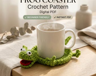 Peut inclure: Un dessous de verre en crochet vert en forme de grenouille avec des yeux blancs et une bouche rouge, supportant une tasse en céramique blanche. Le texte indique : FROG COASTER Crochet Pattern Digital PDF, BEGINNER FRIENDLY, INSTANT PDF.
