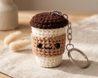 Peut inclure: Un porte-clés en forme de tasse à café crochetée avec un couvercle marron foncé, un corps crème et une manchette marron clair. La manchette présente deux petits yeux en perles noires et une bouche noire brodée. Une chaîne et un anneau en métal argenté sont fixés au sommet.