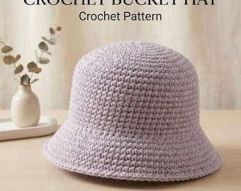 Peut inclure: Un chapeau bob en crochet de couleur violet clair avec un motif de mailles texturé. Le texte CROCHET BUCKET HAT Crochet Pattern est visible en haut de l'image. Le chapeau présente une calotte arrondie et un bord incliné vers le bas.