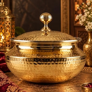 May include: A polished gold-toned metal serving bowl with a matching lid featuring a rounded finial handle. The exterior surface of the bowl and lid displays a distinct hammered texture, creating a reflective finish across the rounded body.