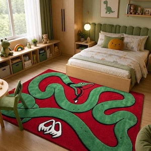 May include: A rectangular area rug featuring a large, winding green snake with a red tongue and white fangs printed on a solid red background. The snake body curves across the entire surface of the rug, creating a bold, graphic pattern.
