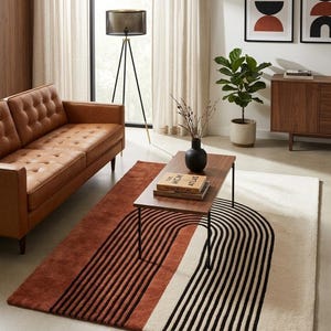 May include: A rectangular area rug featuring a bold graphic design of black concentric arch lines set against a split-color background of deep terracotta orange and cream white, creating a striking visual contrast on the floor surface.