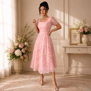 May include: A light pink midi dress featuring a square neckline, short puff sleeves, and a fitted bodice that transitions into a flared skirt. The fabric is adorned with delicate, tonal floral embroidery and appliqué details throughout the garment.