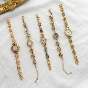 May include: Five gold-tone wristwatches with decorative link bracelets featuring various colorful cabochon stones. Each watch has a unique dial color, including pink, white, and blue, with metallic gold hands and markers on the watch faces.