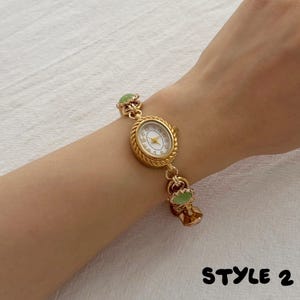 May include: A gold-toned wristwatch with an oval face, white dial, and Arabic numerals. The watch band features a twisted rope-style metal chain interspersed with small, round, light green cabochon accents. The text STYLE 2 appears in black.