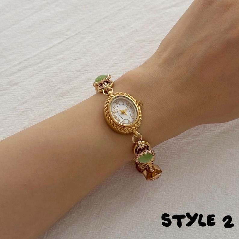 May include: A gold-toned wristwatch with an oval face, white dial, and Arabic numerals. The watch band features a twisted rope-style metal chain interspersed with small, round, light green cabochon accents. The text STYLE 2 appears in black.