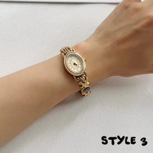 May include: A gold-tone wristwatch featuring an oval-shaped face with a cream dial, black numerals, and a bezel accented with small clear stones. The watch band consists of ornate gold-colored metal links with a dark circular inlay. Text: STYLE 3.