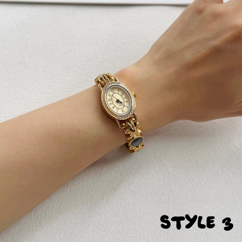 May include: A gold-tone wristwatch featuring an oval-shaped face with a cream dial, black numerals, and a bezel accented with small clear stones. The watch band consists of ornate gold-colored metal links with a dark circular inlay. Text: STYLE 3.
