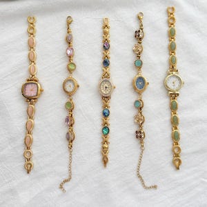 May include: Five gold-tone wristwatches arranged vertically. The first has pink oval stones and a square face. The second features multicolored faceted stones. The third has iridescent circular inlays. The fourth displays floral and blue accents. The fifth has green oval stones.