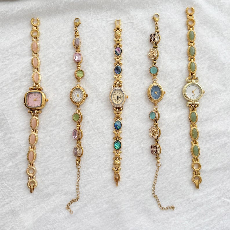 May include: Five gold-tone wristwatches arranged vertically. The first has pink oval stones and a square face. The second features multicolored faceted stones. The third has iridescent circular inlays. The fourth displays floral and blue accents. The fifth has green oval stones.