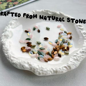 May include: A white ceramic dish with a textured, uneven rim containing a collection of small, marquise-shaped beads in shades of pink, green, brown, and iridescent white, with the text CRAFTED FROM NATURAL STONE printed above the dish.