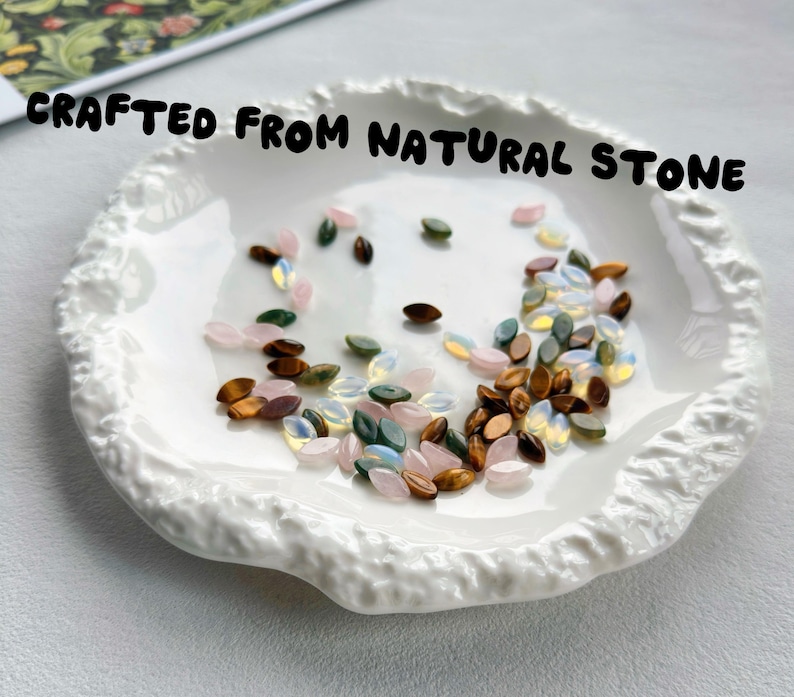 May include: A white ceramic dish with a textured, uneven rim containing a collection of small, marquise-shaped beads in shades of pink, green, brown, and iridescent white, with the text CRAFTED FROM NATURAL STONE printed above the dish.