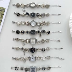 Pode incluir: Oito relógios de pulso com berloques em tom prateado dispostos numa fila vertical. Cada relógio apresenta uma combinação única de berloques pendentes, incluindo estrelas, luas, corações, flores e pérolas, presos a uma corrente de elos.