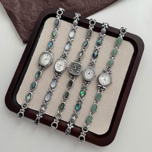 May include: Five silver-toned wristwatches with link bracelets featuring oval inlays of iridescent abalone shell, white mother-of-pearl, and green stone. The watches have varied dial shapes including oval, round, and square with analog displays.