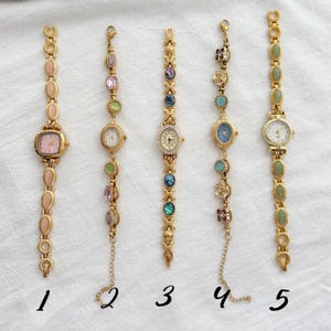 May include: Five gold-toned wristwatches arranged vertically and numbered one through five. Each watch features a unique link bracelet adorned with various colored stones, including pink, green, blue, and iridescent accents, with analog clock faces.
