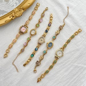 May include: Five gold-tone wristwatches with decorative link bracelets. Each watch features a unique dial color, including pink, white, and blue, accented with various oval and round gemstones or floral-shaped metal charms along the watch bands.