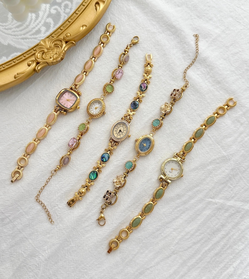 May include: Five gold-tone wristwatches with decorative link bracelets. Each watch features a unique dial color, including pink, white, and blue, accented with various oval and round gemstones or floral-shaped metal charms along the watch bands.