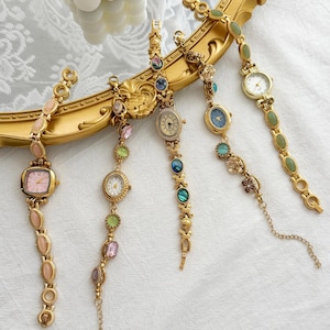 May include: Five gold-tone wristwatches with decorative link bracelets featuring various colored stones and gems. Each watch has a unique face design, including pink, white, and blue dials, arranged in a fan shape over a gold-toned ornate frame.
