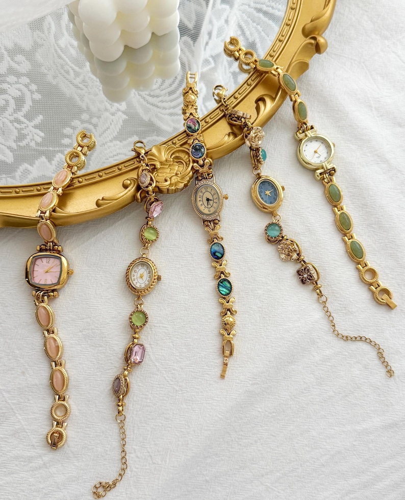 May include: Five gold-tone wristwatches with decorative link bracelets featuring various colored stones and gems. Each watch has a unique face design, including pink, white, and blue dials, arranged in a fan shape over a gold-toned ornate frame.