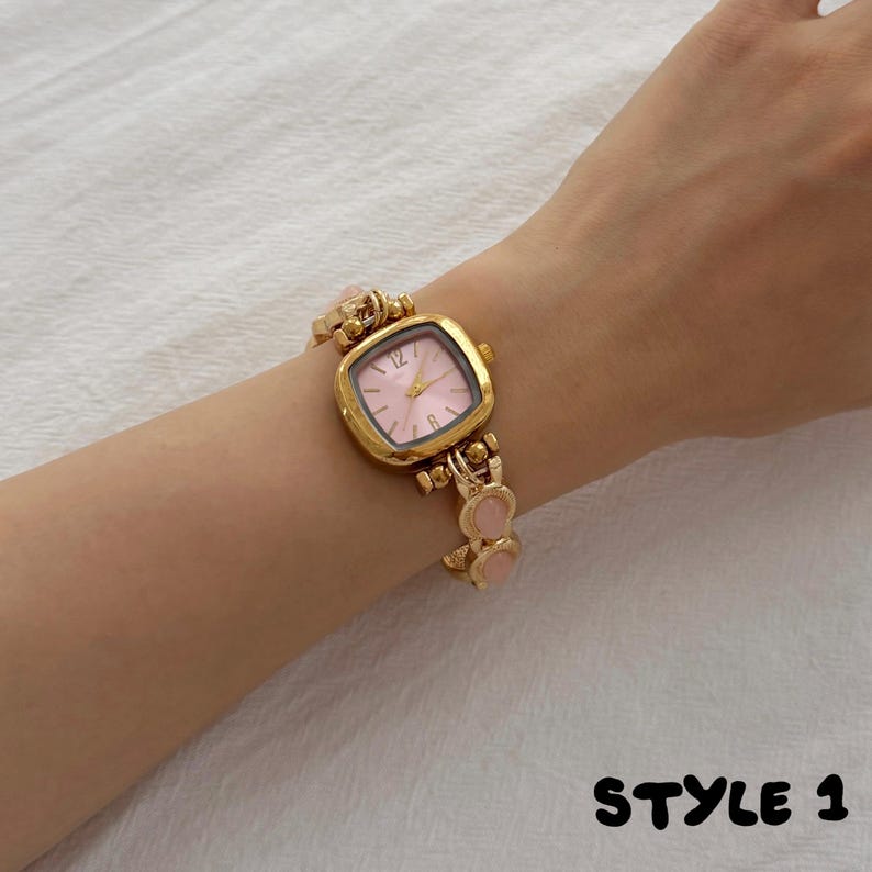 May include: A gold-tone wristwatch featuring a square case with a soft pink dial and gold hands. The watch band is composed of alternating gold-tone metal links and smooth, round pink accents. The text STYLE 1 is visible in the bottom corner.