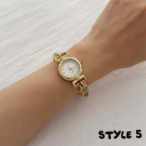 May include: A gold-tone quartz watch featuring a round white face with thin gold hands and markers. The watch band is constructed from gold-tone metal links interspersed with small, smooth, round sage green beads. The text STYLE 5 appears in black.