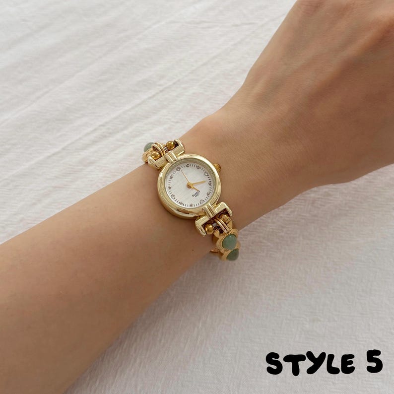 May include: A gold-tone quartz watch featuring a round white face with thin gold hands and markers. The watch band is constructed from gold-tone metal links interspersed with small, smooth, round sage green beads. The text STYLE 5 appears in black.