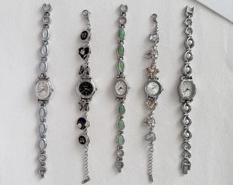 May include: Five silver-tone wristwatches arranged vertically. The first has oval white links, the second features black heart and butterfly charms, the third has oval green stone links, the fourth displays dangling charms, and the fifth has round white pearl-like links.