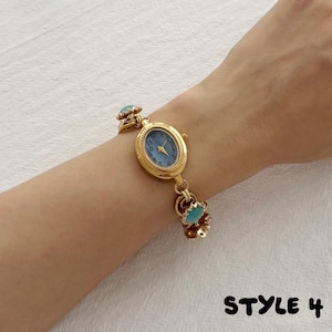 May include: A gold-tone wristwatch with an oval face, a light blue dial, and black Arabic numerals. The watch features a decorative chain-link bracelet accented with small, round, light blue cabochon stones. The text "STYLE 4" is visible.