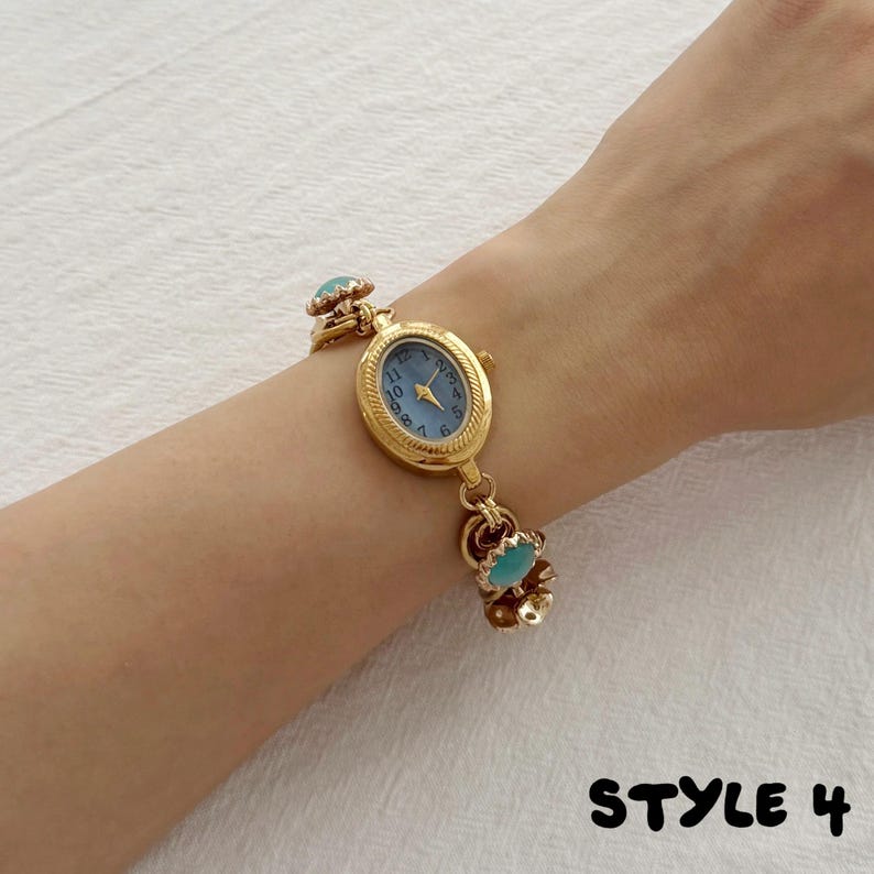 May include: A gold-tone wristwatch with an oval face, a light blue dial, and black Arabic numerals. The watch features a decorative chain-link bracelet accented with small, round, light blue cabochon stones. The text "STYLE 4" is visible.