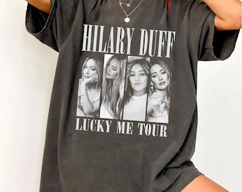 May include: A charcoal gray short-sleeve t-shirt featuring a graphic print with the text HILARY DUFF at the top and LUCKY ME TOUR at the bottom. The center graphic displays four black and white portrait photographs of the same person in a row.