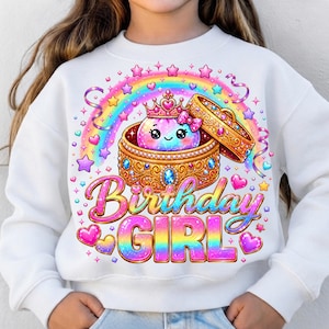 May include: A white sweatshirt featuring a graphic of a smiling, iridescent character wearing a crown inside an ornate gold jewellery box. Above is a rainbow with stars and hearts. The text "Birthday GIRL" appears in large, colourful, gradient letters.