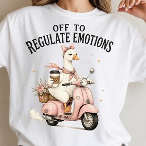 May include: A white t-shirt featuring a graphic of a white goose wearing a pink bow and scarf, riding a pink scooter while holding a coffee cup. The text above the goose reads OFF TO REGULATE EMOTIONS. A basket of flowers is attached to the back.