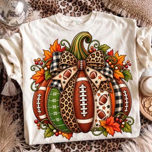 May include: A cream-colored t-shirt featuring a large graphic of a pumpkin designed with football elements. The pumpkin includes a green field yard line pattern, brown football textures, leopard print, and a black and white plaid bow.