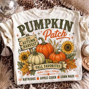 May include: A cream-colored t-shirt featuring a graphic with the text PUMPKIN Patch, EST. AUTUMN HARVEST, FALL FAVORITES, Gather, Enjoy, Celebrate, HAYRIDES, APPLE CIDER, and CORN MAZE, alongside illustrations of pumpkins, sunflowers, and leaves.