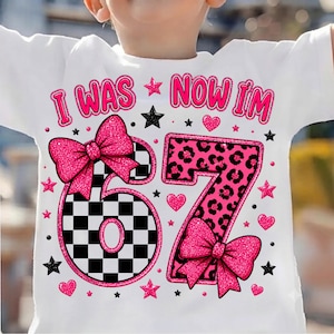 May include: A white t-shirt featuring the text I WAS NOW I'M in pink, with the number 67. The number 6 is black and white checkered, and the number 7 has a pink leopard print. Both numbers are decorated with large, glittery pink bows and stars.