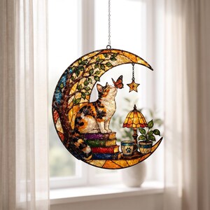 May include: A crescent moon-shaped stained glass suncatcher featuring a calico cat sitting on a stack of books, interacting with a butterfly. The design includes a tree, a small lamp, a mug with a paw print, a potted plant, and a hanging star charm.