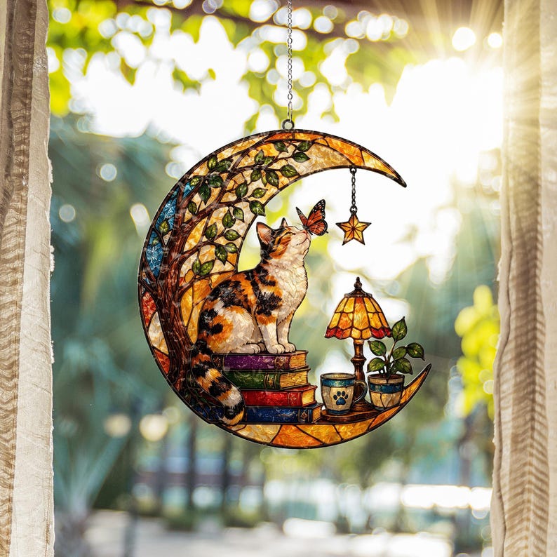 May include: A crescent moon-shaped stained glass suncatcher featuring a calico cat sitting on a stack of books, interacting with a butterfly. The design includes a small hanging star, a table lamp, a potted plant, and a mug with a paw print design.