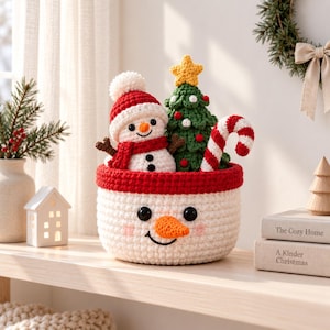 May include: A white crocheted basket with a snowman face, containing a small crocheted snowman wearing a red hat, a green crocheted Christmas tree with a yellow star, and a red and white striped candy cane. The basket has a red rim.