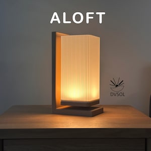 May include: A table lamp with a dark brown L-shaped base and a rectangular, vertically ribbed, translucent white shade that emits a warm, soft glow. The word ALOFT is printed in white above the lamp, and a DVSOL logo is visible to the right.