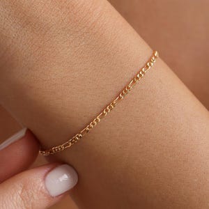 May include: A delicate gold chain bracelet featuring a classic figaro link design. The bracelet consists of a repeating pattern of one elongated oval link followed by three smaller circular links, creating a refined and continuous metallic texture.