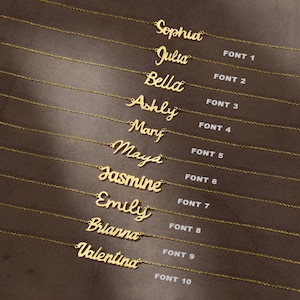 May include: A collection of ten gold-colored name necklaces displayed in a vertical arrangement. Each necklace features a different cursive script font, labeled from FONT 1 to FONT 10, showcasing names including Sophia, Julia, Bella, Ashly, Mary, Maya, Jasmine, Emily, Brianna, and Valentina.